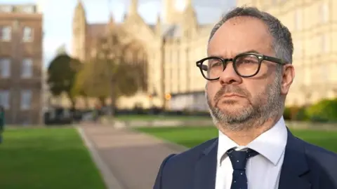 BBC Paul Scully, a man wearing glasses, a blue suit and white shirt