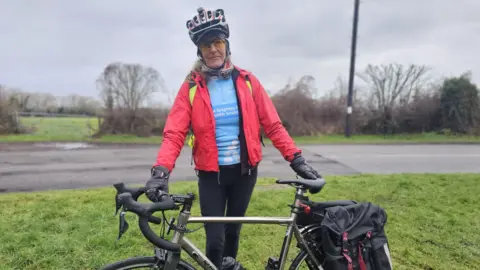Dawn is wearing a silver and red helmet and yellow cycling glasses. She has a blue jersey on with a red waterproof jacket and black leggings. She is standing behind her bike on a grass verge. 