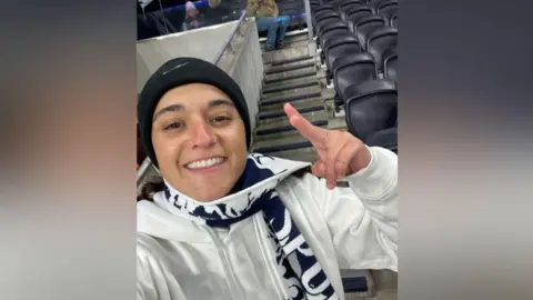 Mandy Damari Emily Damari, a young woman wearing a Spurs scarf and a black beanie hat, smiles in the stands of a football ground