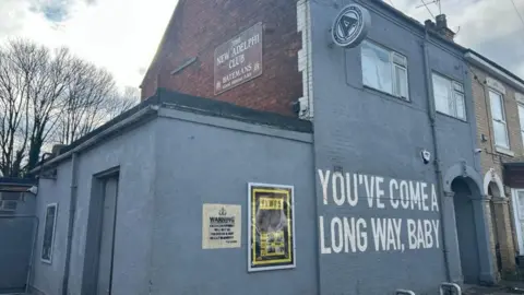 Charlie O'Loughlin/BBC Eksterior Klub Adelphi yang baru. Sepatu foto itu bangunan yang dicat abu-abu dengan bagian atas bata merah. Mural besar di dinding berbunyi, "Anda sudah jauh, sayang." Ada plakat peringatan dan poster berbingkai di dekat pintu masuk.