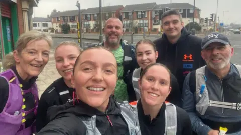 A group of runners male and female in running kit pause for a street selfie. Grant Williams is rear centre left, with a pale green t-shirt and black tracksuit top.