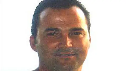 Essex Police Stuart Campbell, who is tanned and has slicked back hair that is black. He has stubble and is smiling at the camera, in front of a white background