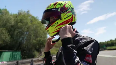 Noah puts on his racing helmet, which is yellow with red and black patterns.