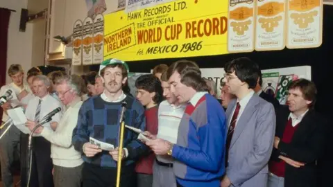 Getty Images Northern Ireland football team members, including Gerry Armstrong in green and white cap dark hair and moustache and checked jumper. Manager Billy Bingham is in a white jumper and glasses. Pat Jennings is in blue, grey and red jumper.