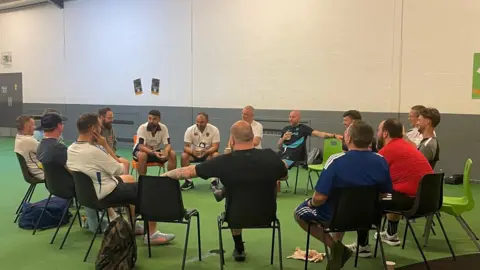 A group of people sit in a circle on chairs inside a sports facility, taking part in a discussion or meeting in a large open indoor space.
