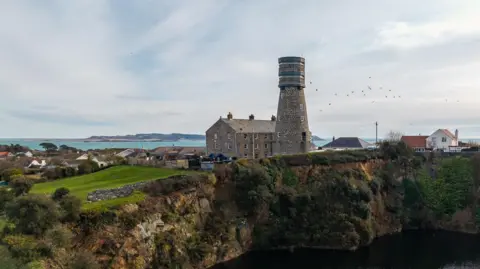 The picture shows a stone tower standing on the edge of a deep, water‑filled quarry, surrounded by greenery and a cluster of nearby houses. The tower appears weathered and historic, with scaffolding or protective covering around its upper section. The quarry below forms a dark, still pool, with steep rocky sides and patches of vegetation growing along the edges.
