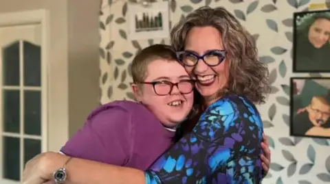 A woman dressed in a blue, and purple top, with shoulder-length dark hair and glasses has a large grin on her her face, as she hugs her son, who is wearing glasses and a purple T-shirt.