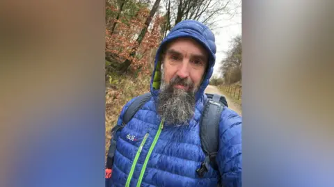 A bearded man in a blue puffa jacket with the hood up looks at the camera. He is wearing a backpack on a woodland path