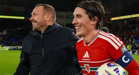 Craig Bellamy and Harry Wilson holding a ball both smile on the field after winning 7-1 against North Macedonia