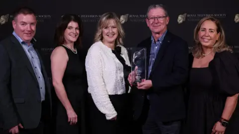 Five people are standing together smiling. Included is Environmental correspondent Louise Cullen, Newsline presenter Tara Mills, Damien Magee (TV news editor),holding an award and TV director Cara. 