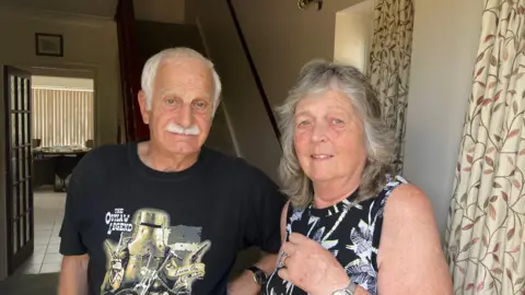 Alexander Stevanovic/BBC Sheila is standing in her front doorway next to her husband Mick. She wears a black and white patterned top, and Mick wears a black T-shirt. He has white hair and a white moustache. Behind them are floral curtains and a staircase.