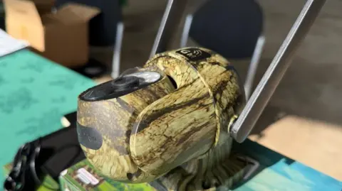 Office for the Leicestershire and Rutland police and crime commissioner A camouflaged camera with two prongs sticking out the back of it. It is on a table. 