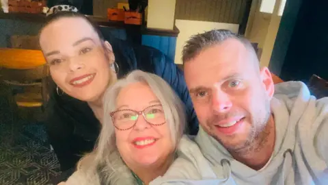 LDRS Campbell pictured with his mum and sister. Campbell is wearing a grey hoodie and holding the camera with both hands. Mallice, in the centre, has long white hair and is wearing burgundy coloured glasses. Campbell's sister, Samantha, has slicked back dark hair and is wearing a black coat.