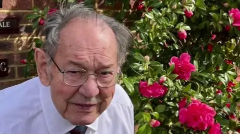 An elderly man with mid-length grey hair is standing in front of pink flowers in a garden wearing a white shirt and tie.