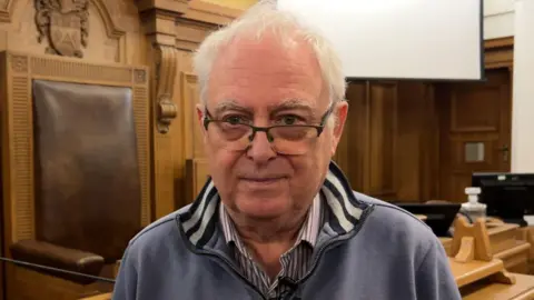 Labour councillor Steve Hey in the council chamber. He is wearing a zip up jumper with a shirt underneath and wears glasses.