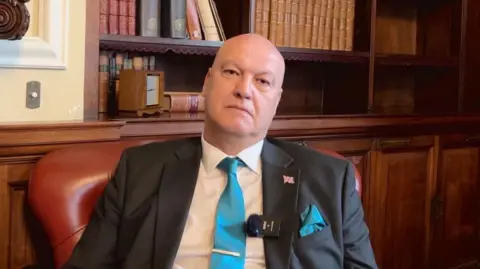BBC Ian Cooper sits in a maroon leather chair, with wooden panelling and a recessed bookcase behind him. He is wearing a white shirt, dark grey suit jacket, with a union flag pin badge and a clip microphone on the lapel, and a teal blue tie. He is looking into the camera with a serious expression.
