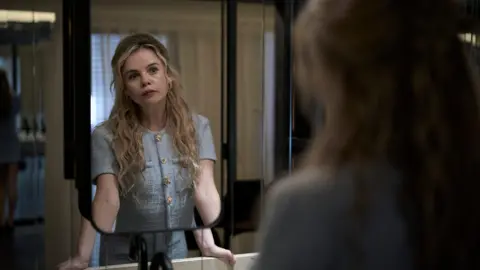 BBC/Left Bank Pictures/James Stack A woman with long blonde hair and wearing a blue buttoned top looks at herself in a bathroom mirror.