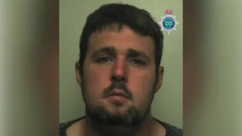 Robert Gillan, a man with dark brown hair and a beard, is standing against a light-coloured backdrop. There is a Staffordshire Police emblem in the top right hand corner of the image.