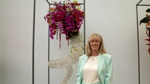 Jonathan Swingler/BBC Nikki Wright standing in front of a display which is made out of purple and pink flowers inside a curved, white structure. She is smiling and has blonde, shoulder-length hair and a full fringe and is wearing a turquoise suit with a white top underneath. There is a white wall behind the display.