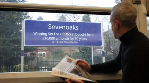 National Lottery A man with short, white hair in a dark jumper reading a newspaper is looking out the train window towards a navy sign. It says "Sevenoaks. Winning Set For Life ticket bought here!".