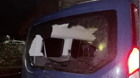 The rear of a blue Ford Tourneo Connect minivan. The windscreen has been smashed and there are several dents in the vehicle. The red brake lights are on. It is dark outside. 