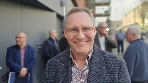 A man with glasses, a paisley shirt and a tweed coat.