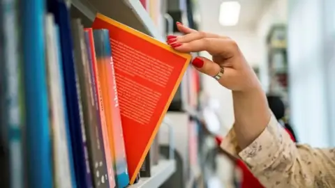 A woman's right hand removing a book from a shelf. The hand has red painted fingernails and a silver ring is being worn on her thumb.