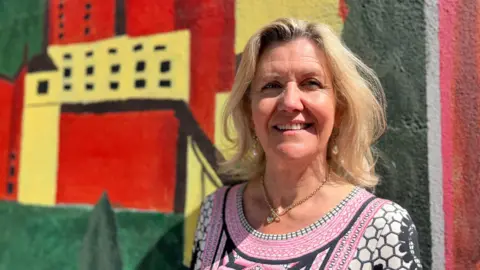 Sue Dougan/BBC A woman stands in front of a colourful mural. She has blonde hair and is wearing a pink, black and white dress.