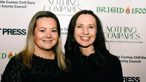 Kildare County Council Katie Lane and Paula O'Brien posing together for an photo at a press event. Katie has long straight blonde hair and Paula has long, straight dark hair. They are both smiling and wearing black dresses. 