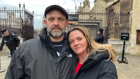 A man and a woman looking at the camera in the foreground with a security guard, black railings and The Houses of Parliament in the background