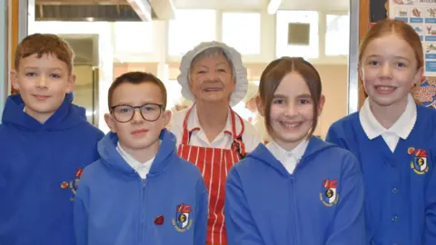 Blaenau Gwent Council Sonia Blachard in catering hat and white uniform with red and white striped apron on top. She is flanked by four pupils in blue uniform hoodies or cardigans and white polo shirts.