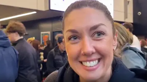 A woman with tied back brown hair smiles at the camera as she stands in a crowd of people at Leeds Station
