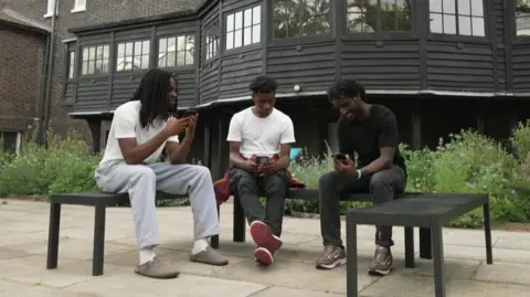 Prince, Henry and Adnan are sitting on some benches, in the gardens of the Museum of the Home. They are casually dressed and smiling as they chat and share some content on their mobile phones. 