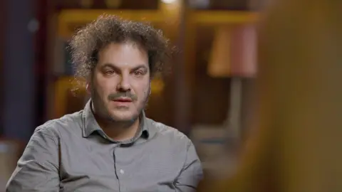 A man with dark curly hair and an eyebrow piercing. He is wearing a grey-blue shirt.