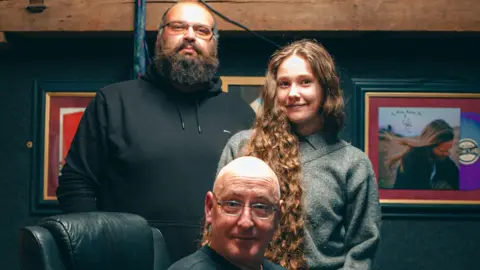 Moose Records/James Revitt Moose Records James Revitt, Macy O and Anthony Shorter (L-R). James has a big black beard and wears a black hoodie, Shorter has a shaved head and glasses while Macy has long wavy brown hair past her shoulders and wears a grey top.
