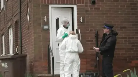 BBC Policeman in black uniform and cap speaks to two forensic officers in white scrubs as they stand outside the entrance to a brick house.