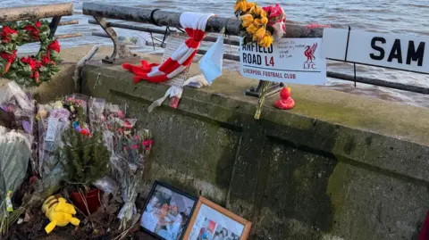 Jasmine Lowe/BBC News A railing next to a large body of water. Attached are lots of bouquets of flowers, posters, messages and photos. The memorial is addressed to Sam and there is his football club Liverpool FC scarfs and merchandise there too. 