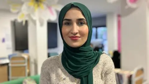 Tara Miran is standing in a room, with a trolley of books behind her. The room is well lit. She has a slight smile and is wearing make up. Her hair and neck are covered by a dark green scarf, and she is wearing a cream knitted jumper. 