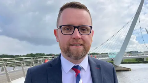 Gary looking at the camera. He is wearing a navy blazer, white shirt, a red, white and blue tie and glasses. He has brown hair and a beard.