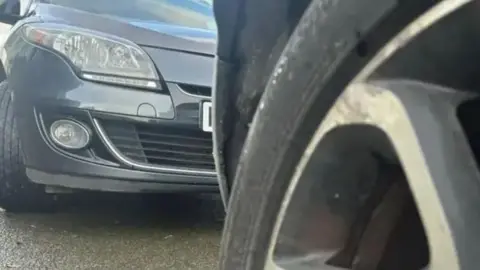 BBC A close-up of two cars parked on a street. The shot has been taken from a low angle. The tyre of one car, which is all that can be seen of it, is in the foreground on the right. The bonnet of another car can be seen on the left of the picture, inches behind the first car.