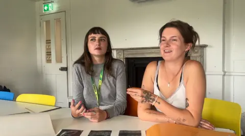 Lauren White and Amelia Watson sitting side-by-side at a white table in a brightly lit room.