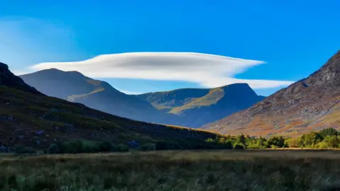 Chris Lawley The image shows a UFO cloud over the Glyderau mountain range in Snowdonia. There is a grass plain in the foreground, with mountains behind and a bright blue sky beyond. 
