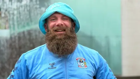 A smiling Lee Worrall wears a Sky Blues hat and top with the club's crest on. He has a long beard. 