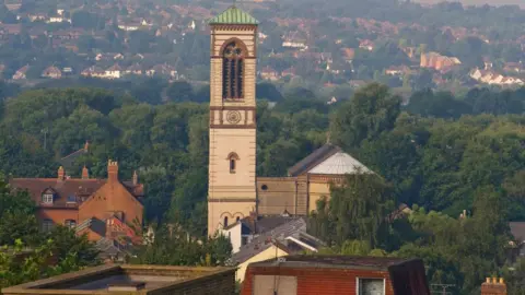 St Barnabas Church A view of the tower of St Barnabas Church among the residential buildings on a clear day. It is surrounded by greenery.