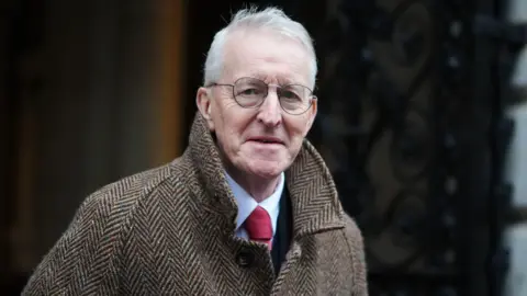 PA Media Hilary Benn walks out of a cabinet meeting at 10 Downing Street. He is an older man with short, white hair. He is wearing a pair of circular glasses, a brown tweet coat and a white shirt with a red tie. He is looking at the camera with a neutral expression on his face.
