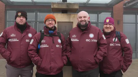 Four men standing in a line. They are all wearing maroon waterproof jackets with the acorns children's hospice logo on the front, as well as the Aston villa foundation logo. Three of the men have beanie hats on, and one has a bald head.