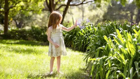 Instagram/Meghan, Duchess of Sussex Lilibet in a summer dress and bare feet in a garden touching a purple flowe