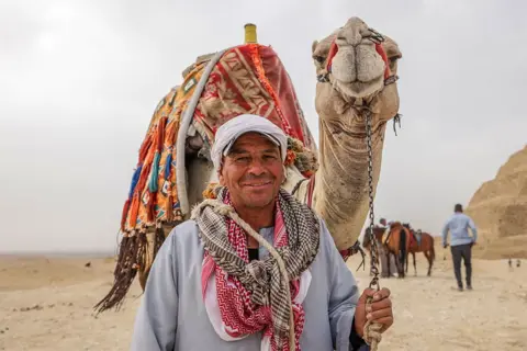 NURETTIN BOYDAK/GETTY IMAGES A man smiling whilst holding on to his camel. There is a pyramid in the background. 