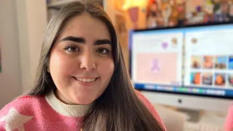 A girl with long dark hair and wearing a pink jumper with white stars on smiles at the camera with a computer screen in soft focus behind her