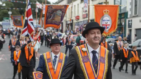 ARCHIVE PHOTO: Orange Order members on parade in Ballynahinch, County Down, in July 2024.  Two men in black bowler hats, dark suits and orange and purple sashes are in the foreground on a wide street.  Other marchers, including some young boys, are marching behind and to the side of them. Some members are holding up colourful Orange Order banners and one is holding a large union flag.